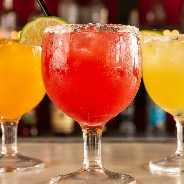Three colorful margaritas, red, orange, and yellow, garnished with lime and salt on a bar.