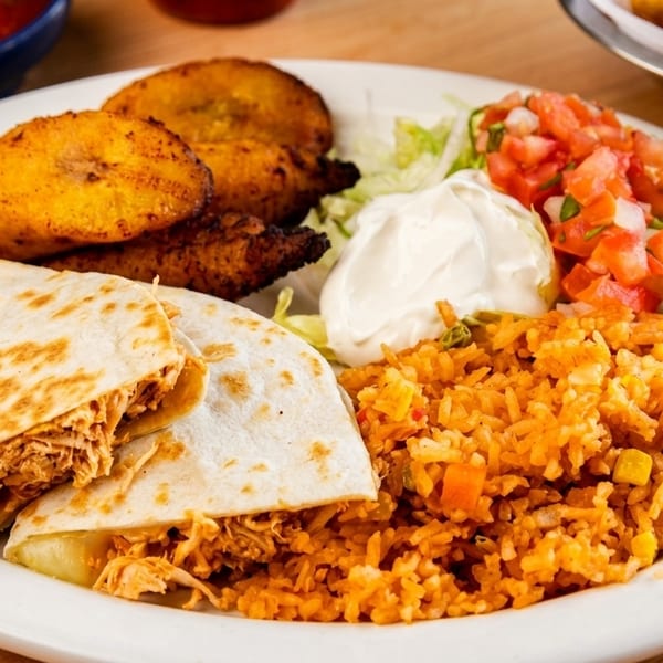 A white plate holds chicken quesadillas, rice, plantains, pico de gallo, and sour cream.