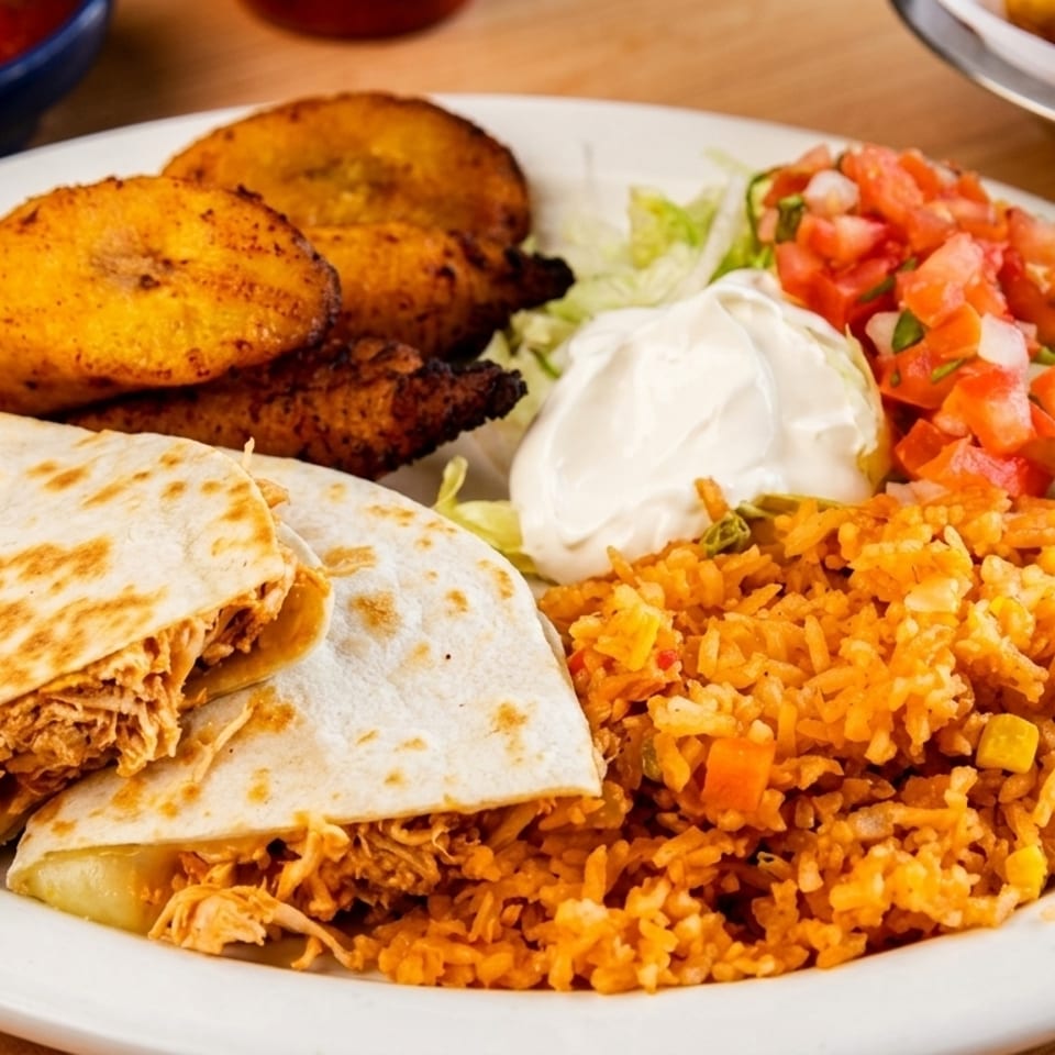 A white plate holds chicken quesadillas, rice, plantains, pico de gallo, and sour cream.