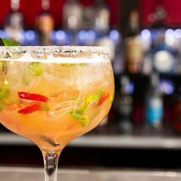 A large margarita with strawberries, mint, and a lime wedge, in front of a blurred bar.