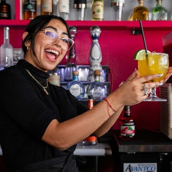 A smiling bartender in glasses holds up a margarita with a lime wedge at a bar.