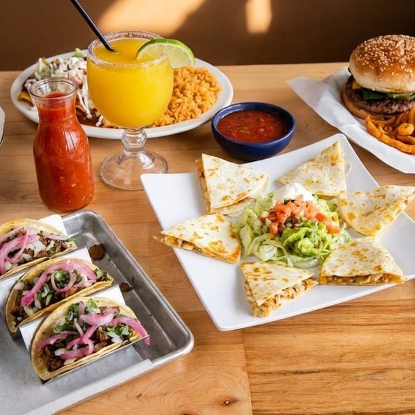 A wooden table laden with various Mexican dishes, including tacos, quesadillas, and a burger.